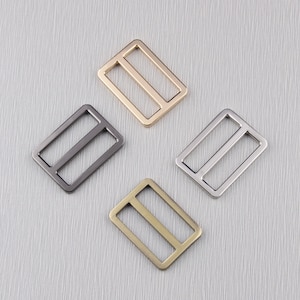 May include: Four metal rectangle sliders in gold, gunmetal, silver, and antique brass. Each slider has two parallel bars inside the rectangular frame. The sliders are arranged on a light grey textured surface.