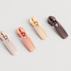 May include: Four metal zipper pulls in various colors: rose gold, gold, bronze, and a muted silver. Each pull has a streamlined design with a curved top and a rectangular base, ideal for sewing projects.