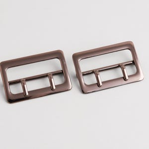 Può includere: Due fibbie in metallo color bronzo. Ogni fibbia ha un telaio rettangolare con tre barre verticali. Le fibbie sono probabilmente destinate a borse, cinture o altri accessori. La finitura è lucida.