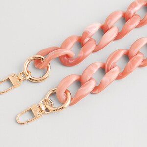 May include: A coral-colored chain strap with gold-tone clasps. The chunky chain links are a marbled peach color. The clasps are a shiny gold color and are attached to the chain.