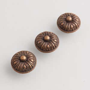 Pode incluir: Três puxadores de armário cor de bronze com um design floral. Cada puxador é redondo com uma esfera central elevada e padrões detalhados em forma de pétala que irradiam para fora. Os puxadores estão dispostos diagonalmente sobre uma superfície branca.