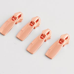 May include: Four rose gold zipper pulls are arranged diagonally on a white surface. Each pull has a rectangular body and a curved top with a hole for the zipper slider. The pulls are made of shiny metal.
