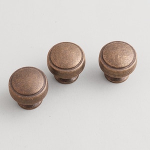 May include: Three antique bronze cabinet knobs. Each knob has a rounded top with a slightly raised edge and a short, cylindrical base. The knobs are a warm, metallic brown color and are arranged on a light gray background.
