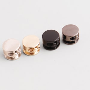 Könnte beinhalten: Vier Metall-Kordelenden in einer Reihe, jeweils mit unterschiedlicher Oberfläche: Roségold, Gold, Schwarz und Gunmetal. Die Stopper sind zylindrisch mit flacher Oberseite und Löchern an den Seiten.
