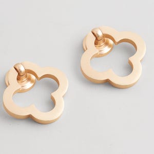 Könnte beinhalten: Zwei goldfarbene Schrankgriffe in Kleeblattform. Jeder Griff hat einen abgerundeten Griff und ein vierblättriges Kleeblatt-Design. Die Hardware besteht aus Metall und ist für eine einfache Installation konzipiert.