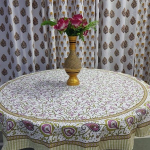May include: A white round tablecloth with a green and pink floral pattern. The tablecloth has a green and white striped border. A vase with pink flowers sits on a table covered with the tablecloth.