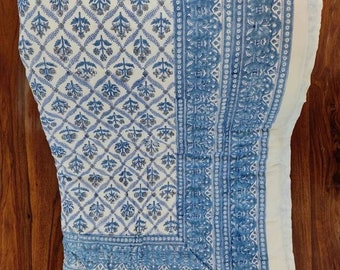 Op de afbeelding: Een gevouwen textiel met een witte achtergrond en een blauw bloemenpatroon. Het ontwerp heeft een herhalend ruitpatroon met gestileerde bloemen en bladeren. Een rand met een vergelijkbaar ontwerp omlijst de rand. Het textiel ligt op een houten oppervlak.