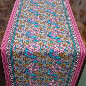 Puede incluir: Un camino de mesa floral con un patrón rosa, azul y dorado sobre un fondo gris. El camino de mesa tiene un borde rosa con un patrón geométrico verde y blanco.