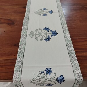 Può includere: Un runner da tavolo bianco con un motivo floreale blu e verde. Il runner ha un bordo decorativo con un motivo floreale.