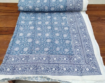 Op de afbeelding: Een blauw-wit bloemmotief quilt, gedeeltelijk opgerold. De quilt heeft een herhalend wit bloemmotief op een blauwe achtergrond. De rand heeft een gedetailleerd, sierlijk patroon in blauw en wit.