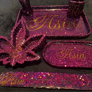 May include: A set of glittery purple resin accessories, including a tray, ashtray, and a cannabis leaf-shaped piece. The name "Asia" is written in gold script on several pieces. The set is designed for use with smoking materials.