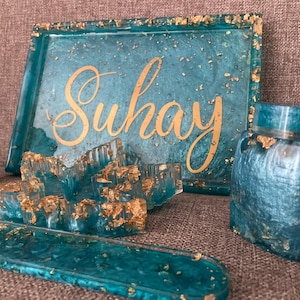 May include: A collection of teal resin home decor items with gold leaf accents. Includes a tray with the name "Suhay" in gold script, a jar, a small sculpture, and a decorative piece. The items are arranged on a neutral surface.