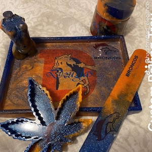 May include: A custom rolling tray set featuring a blue and orange color scheme with glitter accents. The set includes a tray, a small container, a rolling tool, and a leaf-shaped piece. The tray has a Bronco's logo and the words "Broncos" on it. The rolling tool also has the word "Broncos" on it.