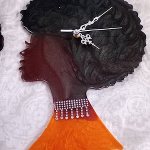 May include: A black and orange resin clock shaped like a woman's profile with an afro hairstyle. The clock face is black with a swirling green pattern and silver clock hands. The woman's neck is adorned with a silver rhinestone necklace.