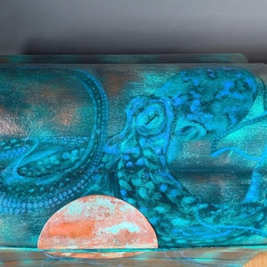 May include: A teal blue wooden box with a raised lid, featuring a painted design of an octopus with a copper coloured sun. The box has a distressed finish.