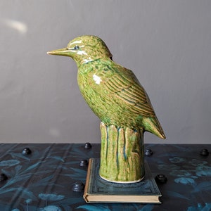 May include: A green ceramic bird ornament with a textured finish, standing on a green base. The bird has a long beak and is placed on a small book. The background includes a gold-framed artwork and a dark blue floral painted trunk.