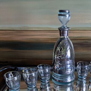 Op de afbeelding: Een set van zes heldere glazen shotglazen met zilveren details en een bijpassende karaf met een zilveren stop. De glazen en de karaf hebben een mat, ge&euml;tst ontwerp van bomen en dieren. De set wordt gepresenteerd op een gespiegelde dienblad.