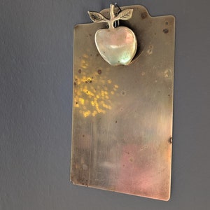 May include: A vintage brass clipboard with an apple-shaped clip. The clipboard is rectangular with rounded corners and a slightly angled bottom edge. The apple clip has a detailed design with leaves.