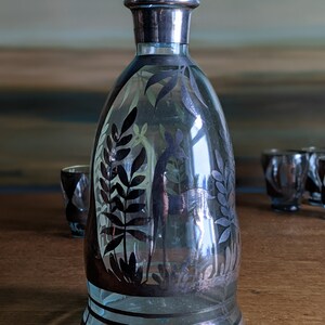 Op de afbeelding: Een heldere glazen karaf met een zilverkleurige bovenkant en een zwart-wit ontwerp van bladeren en een vogel. De karaf staat op een houten tafel met drie kleine shotglazen.