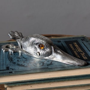 May include: A silver bottle opener in the shape of a bird's head, with an orange eye, placed on a stack of blue books. The opener's beak is extended, and the bird's head has detailed feathering.