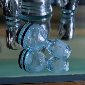 Op de afbeelding: Twee heldere blauwe glazen flessenstoppers met zwarte banden rond de bovenkant. De stoppers liggen op een reflecterend oppervlak.