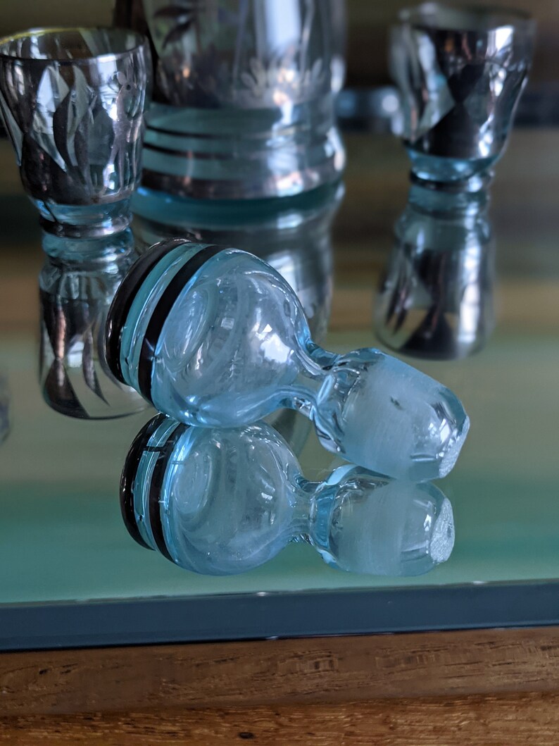 Op de afbeelding: Twee heldere blauwe glazen flessenstoppers met zwarte banden rond de bovenkant. De stoppers liggen op een reflecterend oppervlak.