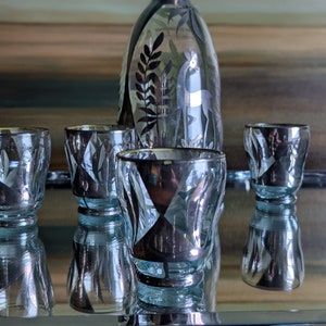 Op de afbeelding: Een set van vier heldere glazen shotglazen met zilveren bladontwerpen en een bijpassende heldere glazen karaf met een zilveren stop en zilveren bladontwerpen.