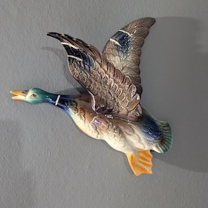 May include: A ceramic wall decoration depicting a duck in flight. The duck features detailed feathering in brown, blue, and white. The head is green with a yellow beak, and the feet are orange. The duck is mounted on a grey wall.