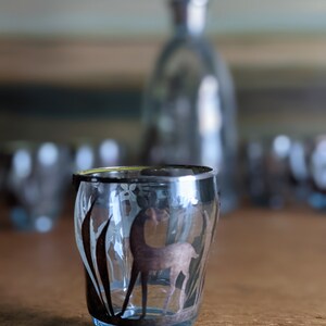 Op de afbeelding: Een helder glazen shotglas met een zilveren rand en een gegraveerd zilveren ontwerp van een hert in een veld. Het glas staat op een houten tafel.