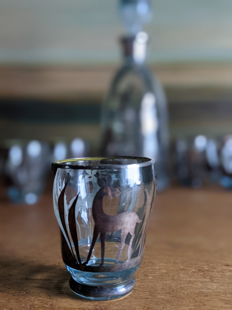 Op de afbeelding: Een helder glazen shotglas met een zilveren rand en een gegraveerd zilveren ontwerp van een hert in een veld. Het glas staat op een houten tafel.