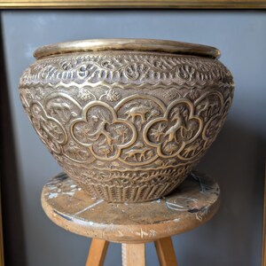 May include: An antique metal bowl with detailed embossed patterns, including animals and floral designs. The bowl is displayed on a small, round wooden stool. The metal has a tarnished appearance, highlighting its age and intricate craftsmanship.