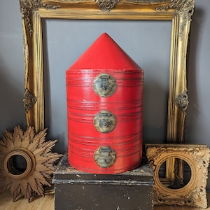 May include: A striking red cylindrical container with a conical top, secured by three brass-toned clasps. The container has horizontal bands and is placed on a black box. Gold-framed decor is in the background. A decorative storage item.