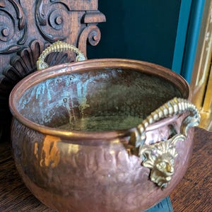 May include: A copper bowl with brass handles, featuring a dragon head design. The bowl has a hammered texture and a patinaed finish. It rests on a stack of blue books. The background includes a dark green wall and wooden carving.