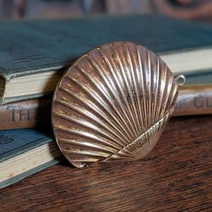 May include: A gold-coloured, seashell-shaped compact. The compact features a detailed, ridged design and a small loop for a chain. It sits on a wooden surface near old books, suggesting a decorative or collectible item.
