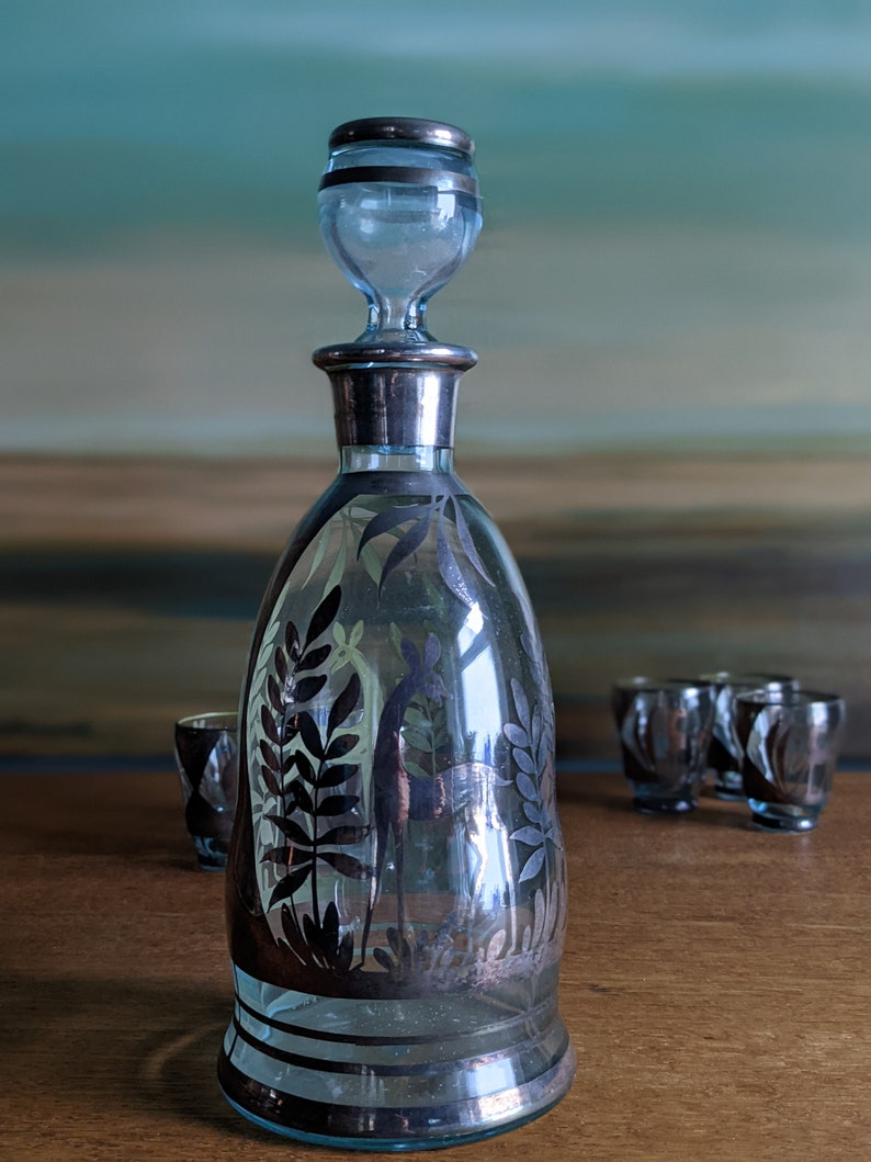 Op de afbeelding: Een heldere glazen karaf met een zilverkleurige bovenkant en een zwart-wit ontwerp van bladeren en een vogel. De karaf staat op een houten tafel met drie kleine shotglazen.