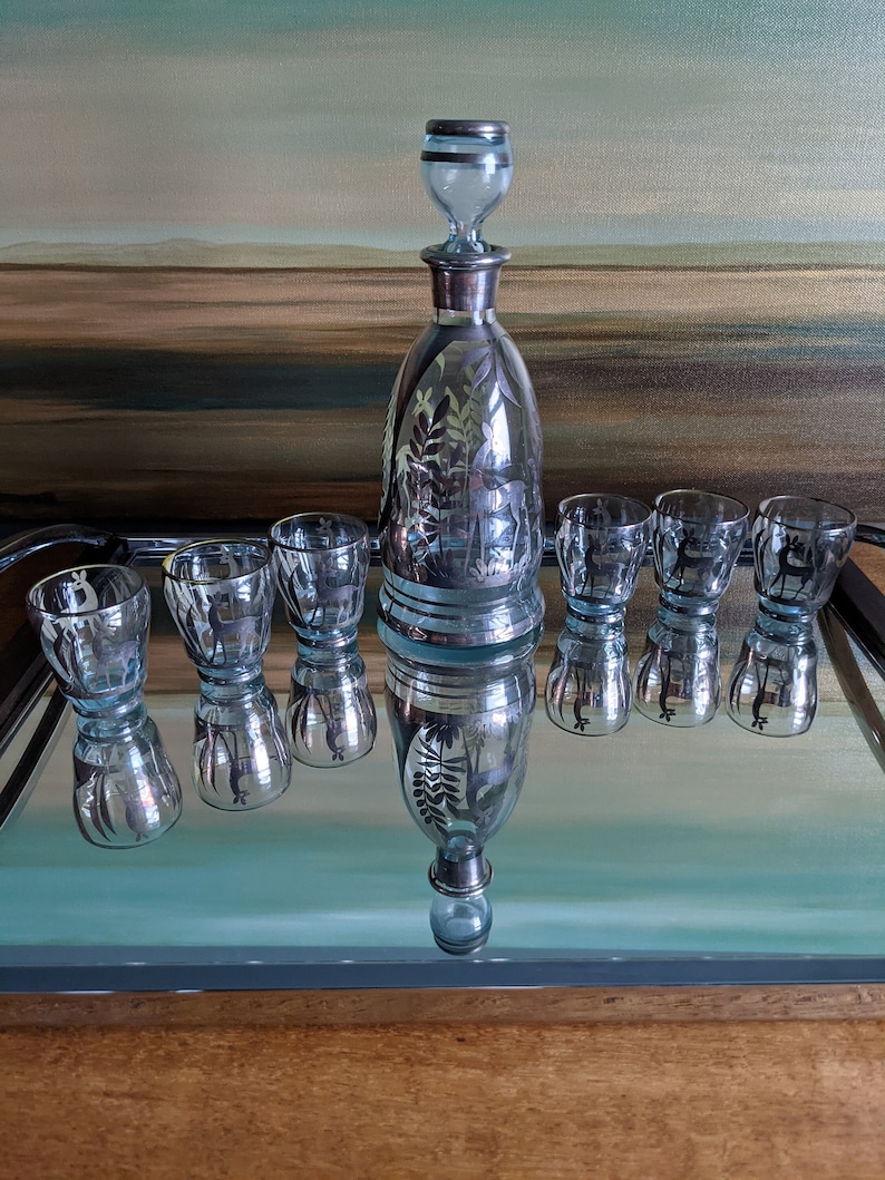 Op de afbeelding: Een set van zes heldere glazen shotglazen met zilveren details en een bijpassende karaf met een zilveren stop. De glazen en de karaf hebben een mat, ge&euml;tst ontwerp van bomen en dieren. De set wordt gepresenteerd op een gespiegelde dienblad.
