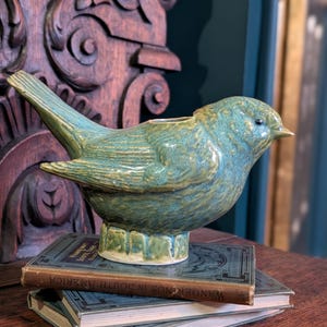 May include: A teal ceramic bird ornament with a textured finish. The bird sits on a stack of vintage books with gold lettering. The books are on a dark wooden surface, with a carved wooden background.
