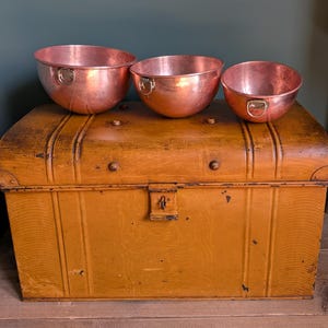May include: Three copper bowls with ring handles sit on a weathered, mustard-coloured trunk. The bowls vary in size, displaying a warm, metallic sheen. The trunk has a vintage aesthetic, with metal accents and a lock.