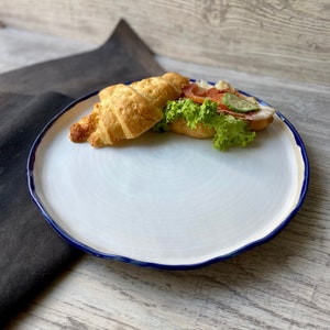 Pode incluir: Um prato de cerâmica branco com uma borda azul, apresentando um sanduíche de croissant com alface, tomate e pepino. O prato está sobre uma superfície de madeira.