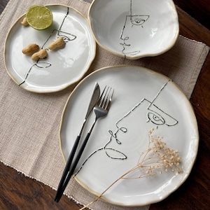 May include: Three white ceramic plates with black line drawings of faces. The plates are on a beige placemat. Two forks and a knife are on the largest plate.