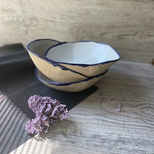 May include: Three ceramic bowls stacked on top of each other. The bowls are a light beige color with a white interior and a dark blue rim.
