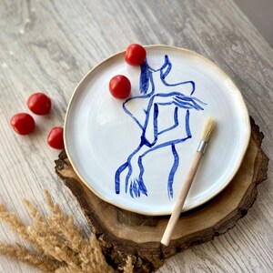 Pode incluir: Um prato de cerâmica branco com um desenho de linha azul de uma pessoa. O prato está sobre uma superfície de madeira com tomates vermelhos e um pincel.