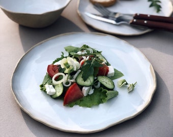 May include: A white ceramic plate with a light brown rim, featuring a salad with fresh ingredients like tomatoes, cucumbers, onions, feta cheese, and leafy greens.