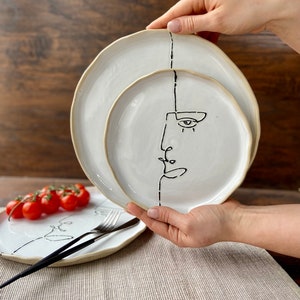 Puede incluir: Dos platos de cerámica blanca con dibujos de caras en líneas negras. El plato más pequeño tiene una cara con un ojo y una nariz. El plato más grande tiene una cara con una línea en el centro.