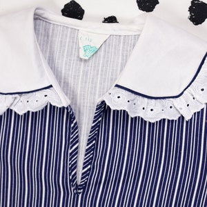 May include: A white collared shirt with a blue and white striped pattern and a white lace trim. The collar has a navy blue trim. The shirt has a small tag that says "C. Rae".