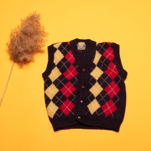 May include: A black, red and cream argyle patterned sleeveless sweater vest with five brown buttons.