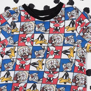 May include: A white t-shirt with a colorful checkered pattern featuring cartoon characters from Looney Tunes. The characters include Bugs Bunny, Daffy Duck, Tweety Bird, and Sylvester the Cat.