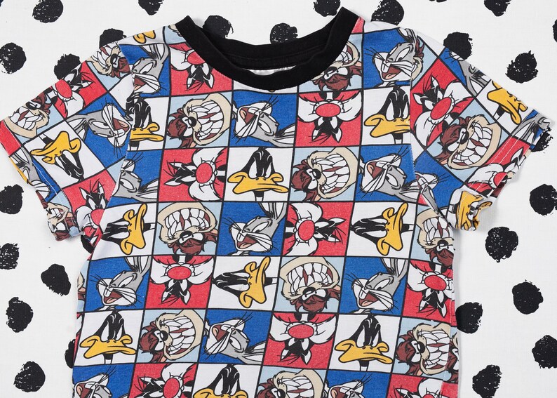 May include: A white t-shirt with a colorful checkered pattern featuring cartoon characters from Looney Tunes. The characters include Bugs Bunny, Daffy Duck, Tweety Bird, and Sylvester the Cat.
