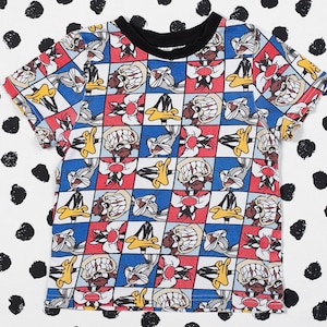 May include: A white t-shirt with a colorful cartoon print of Bugs Bunny and Daffy Duck. The print features a checkered pattern with red, blue, and yellow squares.