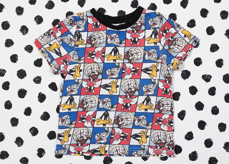 May include: A white t-shirt with a colorful cartoon print of Bugs Bunny and Daffy Duck. The print features a checkered pattern with red, blue, and yellow squares.